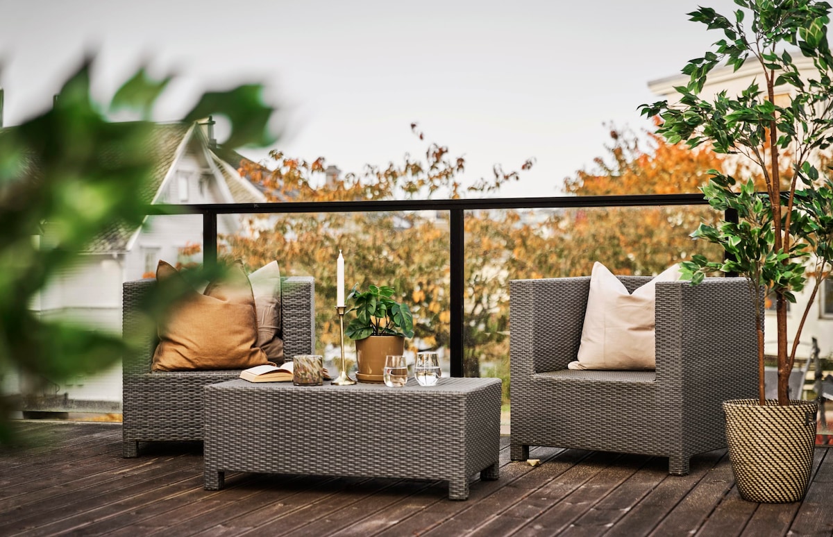 A balcony area features two wicker armchairs, each complemented by soft cushions. A low coffee table sits in between, adorned with decorative items. Lush greenery surrounds the space, providing a serene view of autumnal trees in the background.