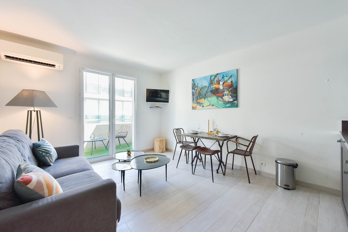 A bright living space is featured, showcasing a grey sofa accompanied by an accent pillow. A dining table for three sits adjacent, while a wall-mounted television is visible. Large glass doors lead to a balcony, enhancing the room's connection to the outdoors.