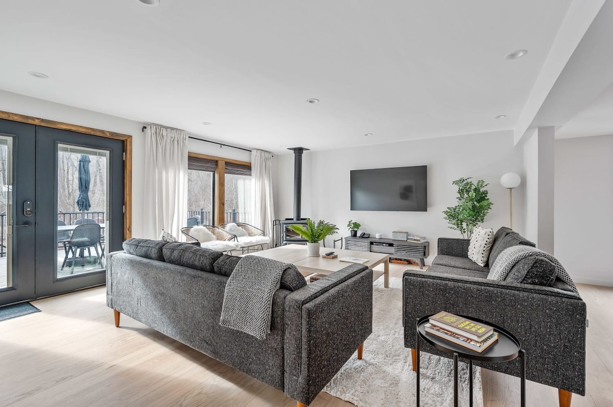 A spacious living room is furnished with dark grey leather sofas and a light area rug. Large windows provide natural light and offer views of the outdoors. A modern entertainment unit holds a flat-screen TV, while a wood-burning fireplace adds warmth to the space.