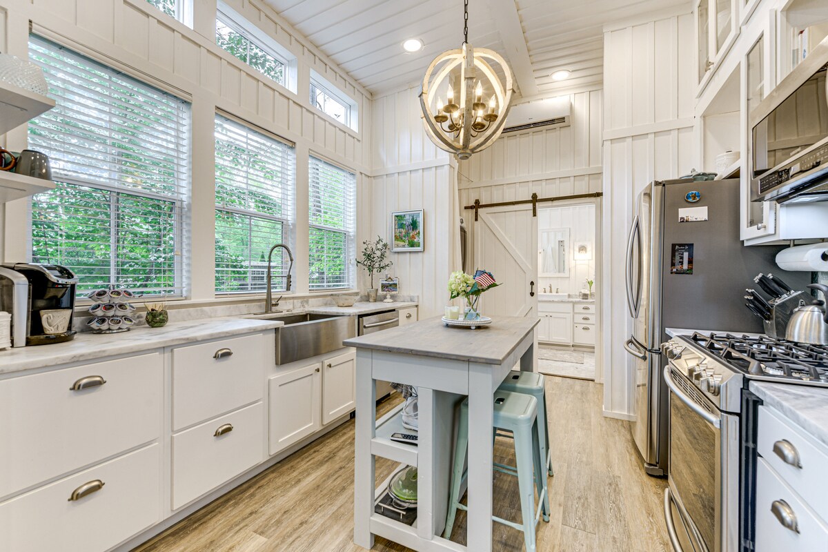 A modern kitchen is showcased, featuring white cabinetry and stainless steel appliances. Large windows fill the space with natural light. A central island with seating is complemented by a chandelier above. This area provides a view of a nearby bathroom, visible through a sliding door.