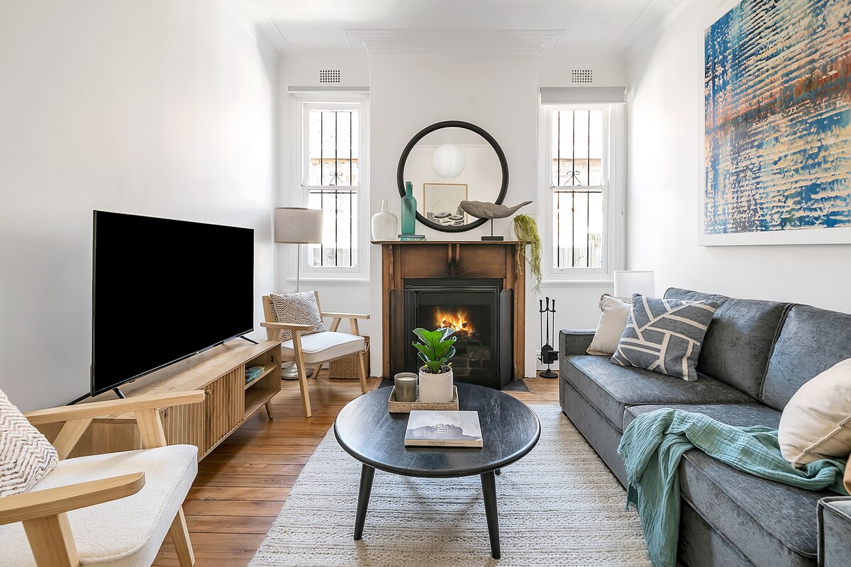 A cozy living area features a comfortable grey sofa, a round coffee table, and a television mounted on the wall. A decorative fireplace adds warmth, surrounded by two large windows adorned with natural light. An artwork adds a splash of color to the space.