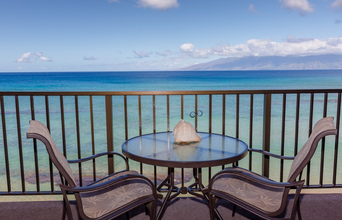 A serene lanai offers a round table with two cushioned chairs, positioned to overlook the ocean. A decorative shell rests on the table, while the expansive sea and distant mountains provide a tranquil backdrop, showcasing varying shades of blue and green in the water.