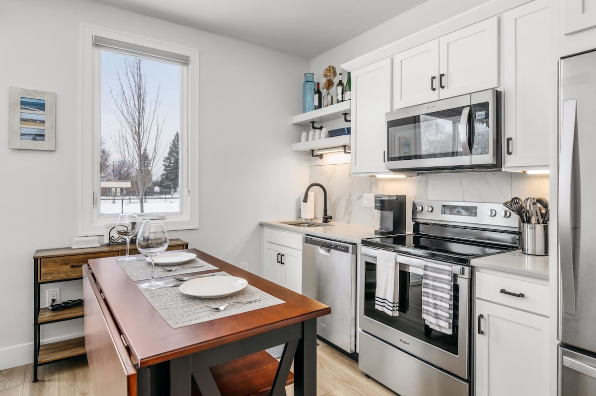 The kitchen showcases modern appliances and a clean design. A wooden dining table is set for two, with glassware and tableware arranged neatly. Natural light enters through a window, highlighting the functional layout and soft color palette.