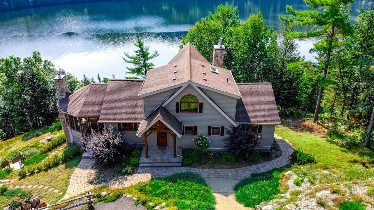 The exterior of the house is shown from an aerial view, surrounded by lush greenery. The property features a welcoming entryway, a pathway made of stone, and a large yard that leads toward the water's edge, where the lake reflects the tree line.