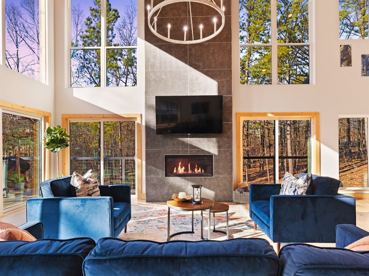 A spacious living area features large floor-to-ceiling windows, allowing natural light to fill the room. Plush blue sofas and accent chairs are arranged around a circular table. A modern gas fireplace and a flat-screen TV create a cozy focal point against a textured wall.