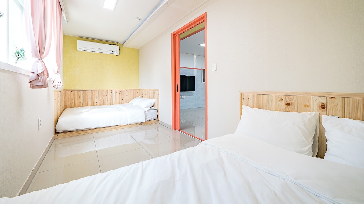 A spacious room is presented with two beds, one positioned on a wooden platform. Soft white linens are neatly arranged on both beds. The walls are painted in pale tones, complemented by a vibrant accent wall. A window allows natural light to brighten the space.