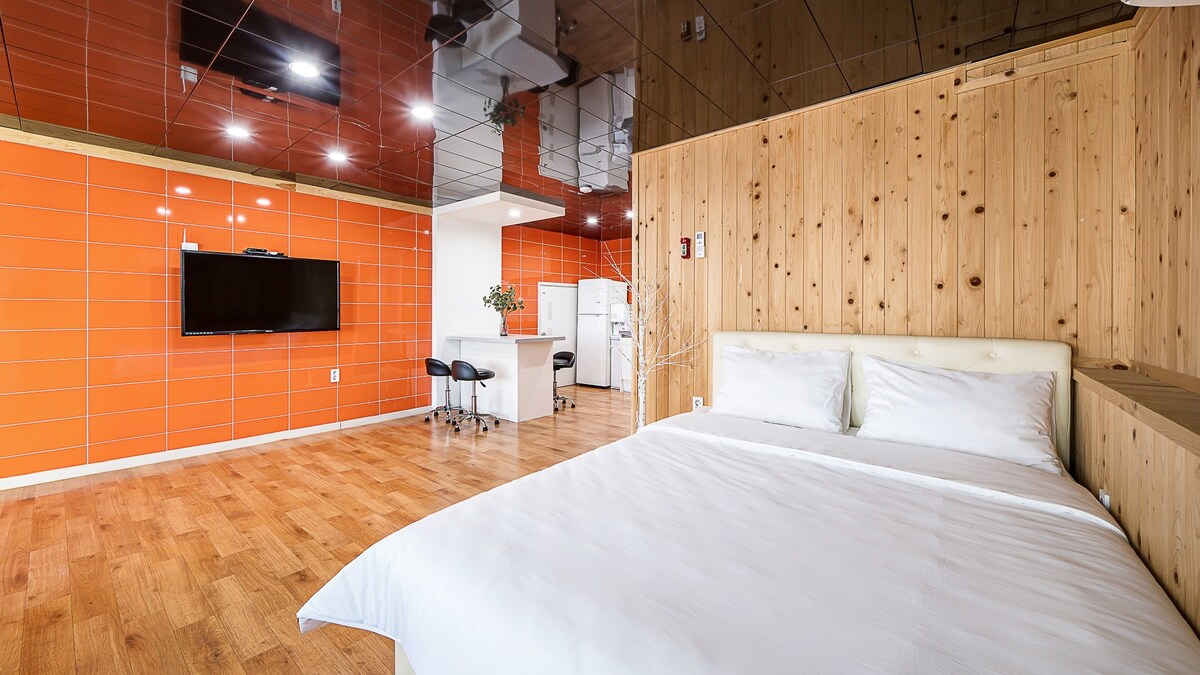 A bright and spacious room features a large, comfortable bed with crisp white linens. A distinctive wood-paneled wall contrasts with the vibrant orange tiles in the background. The area includes a kitchen space with stools and a television mounted on the wall.