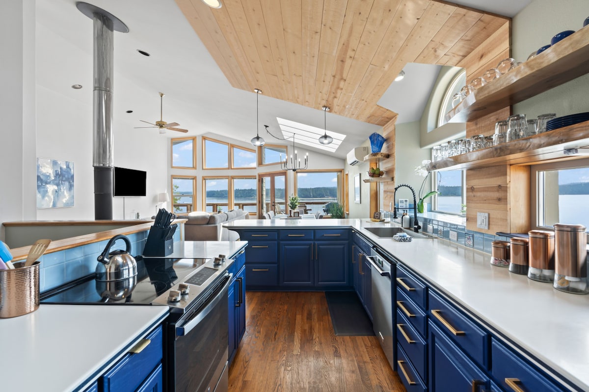 The home’s fully equipped kitchen features navy blue cabinetry and a spacious countertop. Large windows showcase views of the water, and a wood ceiling adds warmth to the design. Stainless steel appliances include an oven and a dishwasher, with glassware and utensils visible on open shelves.
