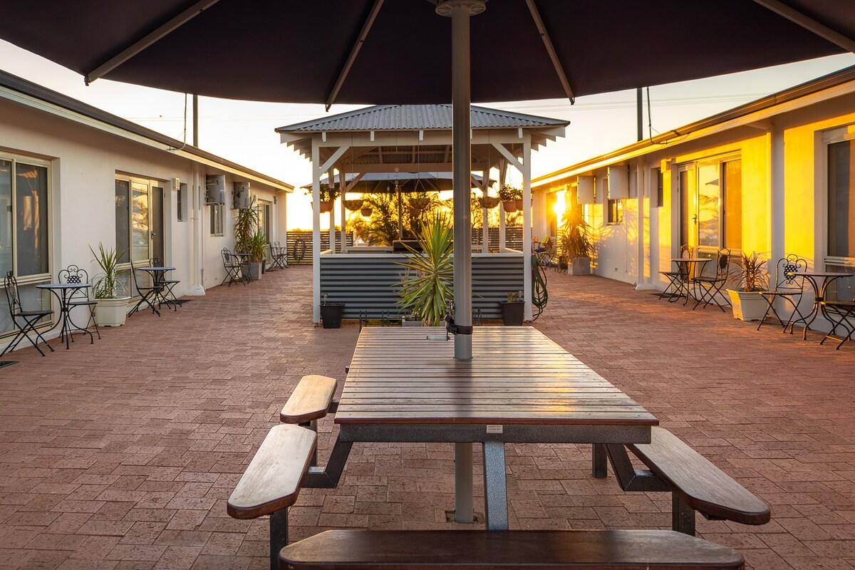 An outdoor communal area features a centrally located wooden table surrounded by benches, providing seating for guests. Two distinct seating areas with wrought iron furniture are visible, while sunset light casts a warm glow over the well-maintained courtyard, enhancing the inviting environment.