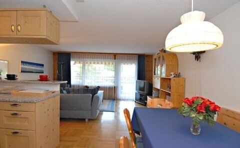 An open-plan living area is visible, featuring a dining table set with a blue tablecloth and fresh flowers. A comfortable sofa is situated across from a television. Natural light fills the space through large windows, and wooden cabinetry complements the interior.