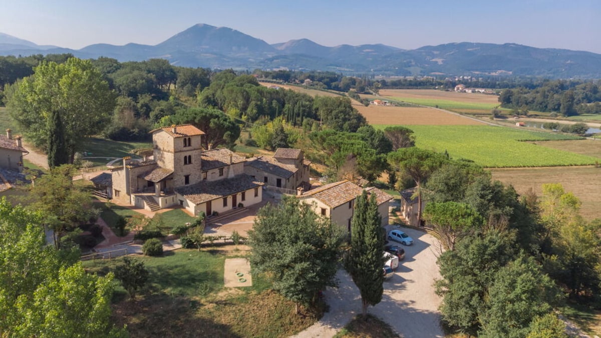Aerial view captures the rustic charm of the Pulciano Apartments, set within an ancient medieval village. Surrounding countryside features rolling hills, vineyards, and lush greenery, enhancing the tranquil atmosphere. Parking spaces are visible, alongside scattered trees that frame the serene landscape.