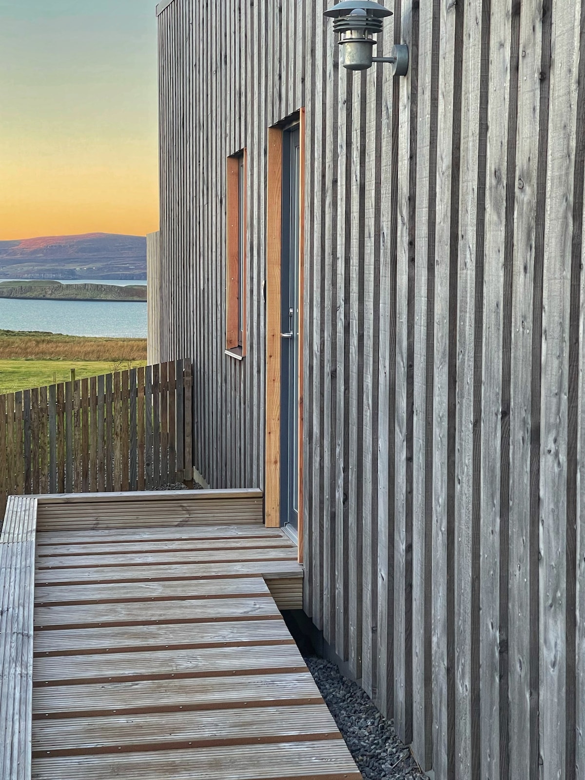 The exterior of a wooden-clad house is shown, featuring a long deck made of wooden planks leading to a door. The surrounding area displays lush greenery and a tranquil body of water in the distance, complemented by soft evening light.