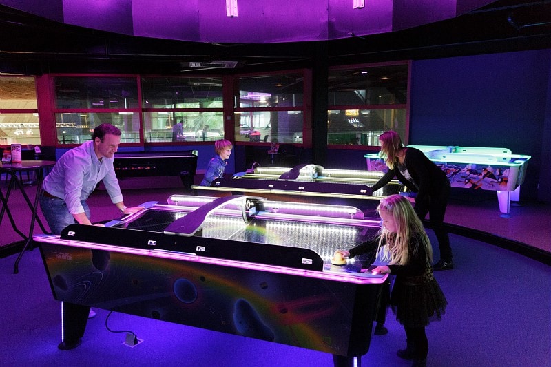 A vibrant indoor space features several air hockey tables illuminated with colorful neon lights. Two adults and two children are engaged in a friendly game, with the table surfaces reflecting the ambient light. Large windows allow glimpses of additional recreational activities in the background.