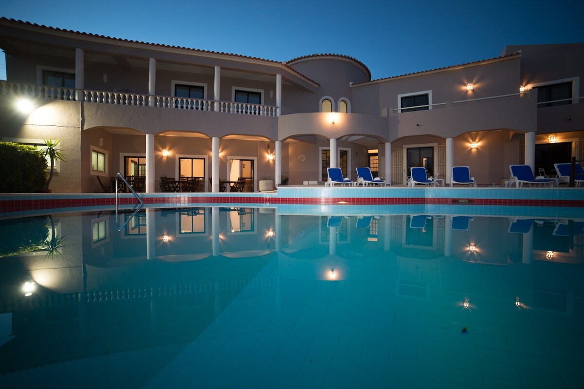 An inviting swimming pool reflects the surrounding multi-level building illuminated at dusk. Lounge chairs are positioned around the pool, and several outdoor seating arrangements are visible on the patio, creating a tranquil environment for relaxation.