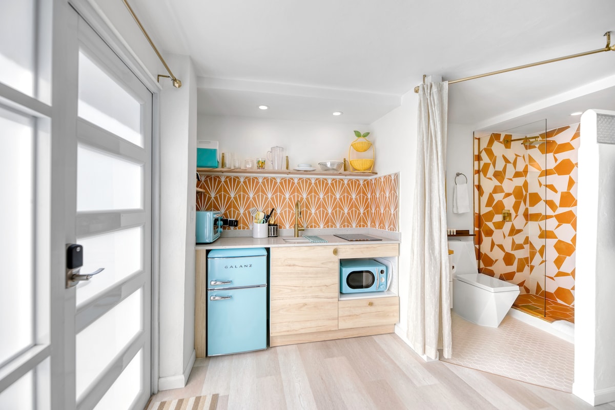 The kitchenette features a colorful backsplash with geometric patterns, sleek wooden cabinetry, and modern appliances. A small refrigerator is prominently displayed alongside a microwave. Natural light illuminates the space, enhancing the bright and airy atmosphere.