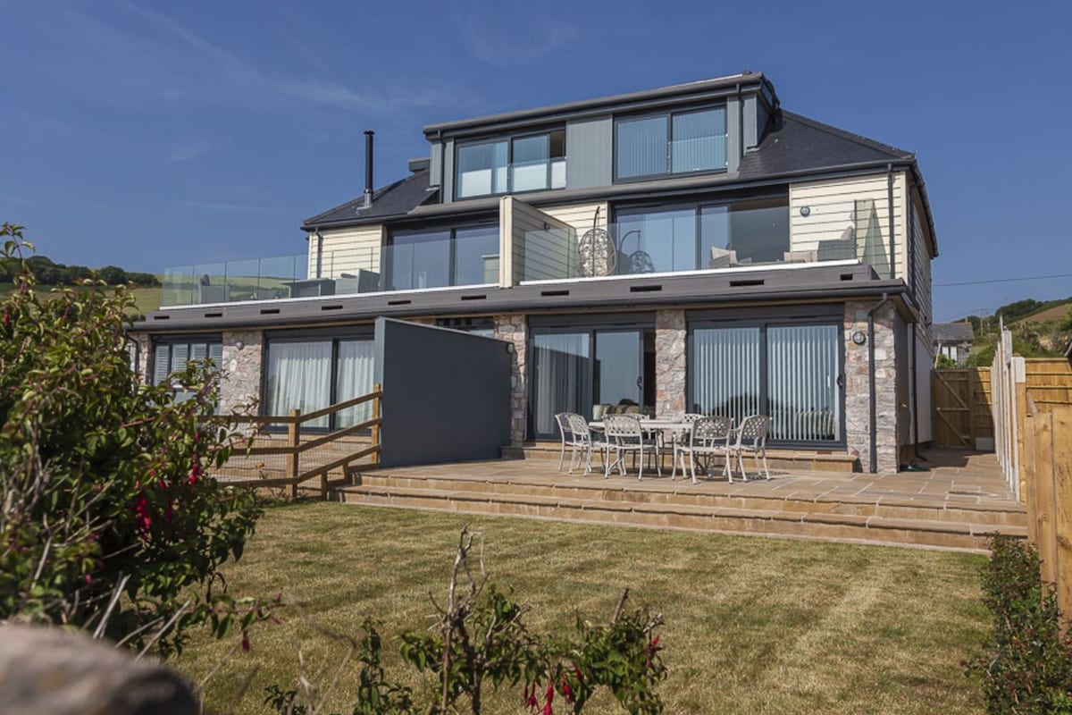 The modern exterior of the holiday home showcases a multi-level structure with large glass doors and balconies. A spacious patio area is equipped with outdoor dining furniture, surrounded by well-kept grass and plants, highlighting a serene coastal setting.