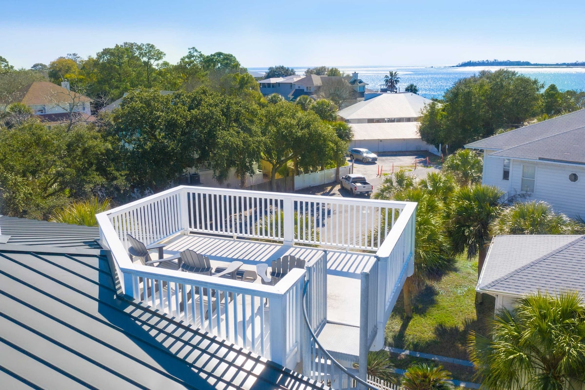 A rooftop terrace offers expansive views of the ocean and surrounding landscape. Lounge chairs are positioned for relaxation, while the railing provides safety without obstructing the scenic vista. The bright sky enhances the coastal ambiance, creating an appealing outdoor retreat.