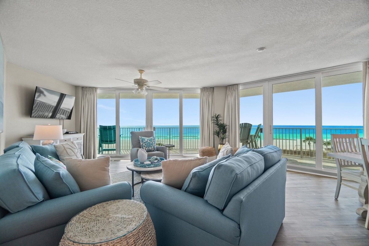 A bright living area features large windows that provide a view of the ocean and beach. Comfortable blue sofas are arranged around a coffee table, with a television mounted on the wall. A dining area is visible, complemented by decor that enhances the seaside setting.