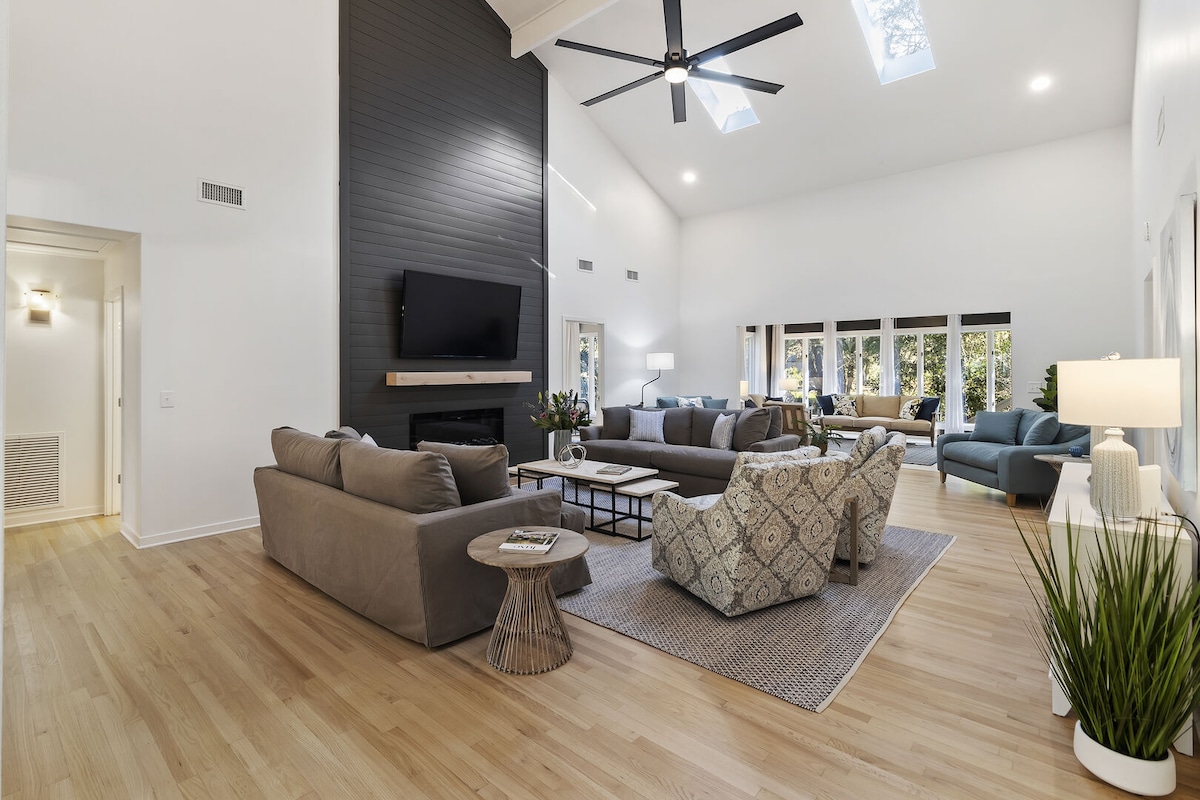 A spacious living area is highlighted by a high ceiling and large windows allowing natural light to illuminate the room. Comfortable seating arrangements include a sectional sofa and armchairs, complemented by a neutral color palette. A television is mounted above a modern fireplace, enhancing the inviting atmosphere.