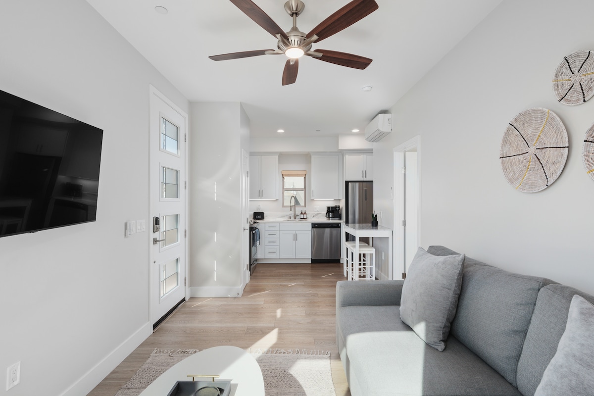 A modern living space is showcased with a comfortable gray sofa and a round coffee table. A flat-screen TV is mounted on the wall, while bright natural light fills the area through large windows. The kitchen is visible in the background, featuring sleek cabinetry and stainless steel appliances.