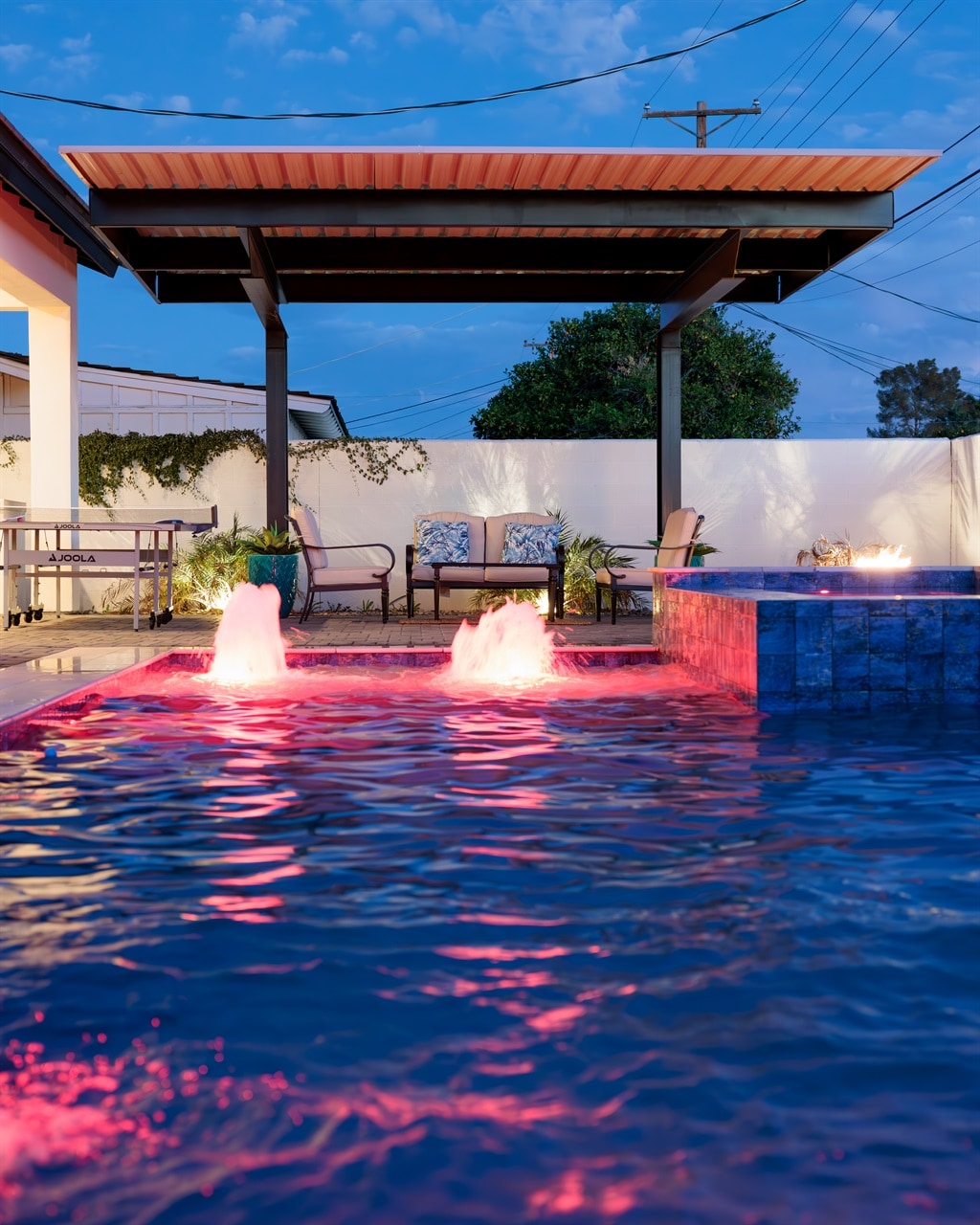 A private heated pool is illuminated with pink water features, creating a relaxing ambiance. Adjacent to the pool, a shaded seating area is furnished with two chairs and is surrounded by lush greenery, providing a serene outdoor space.