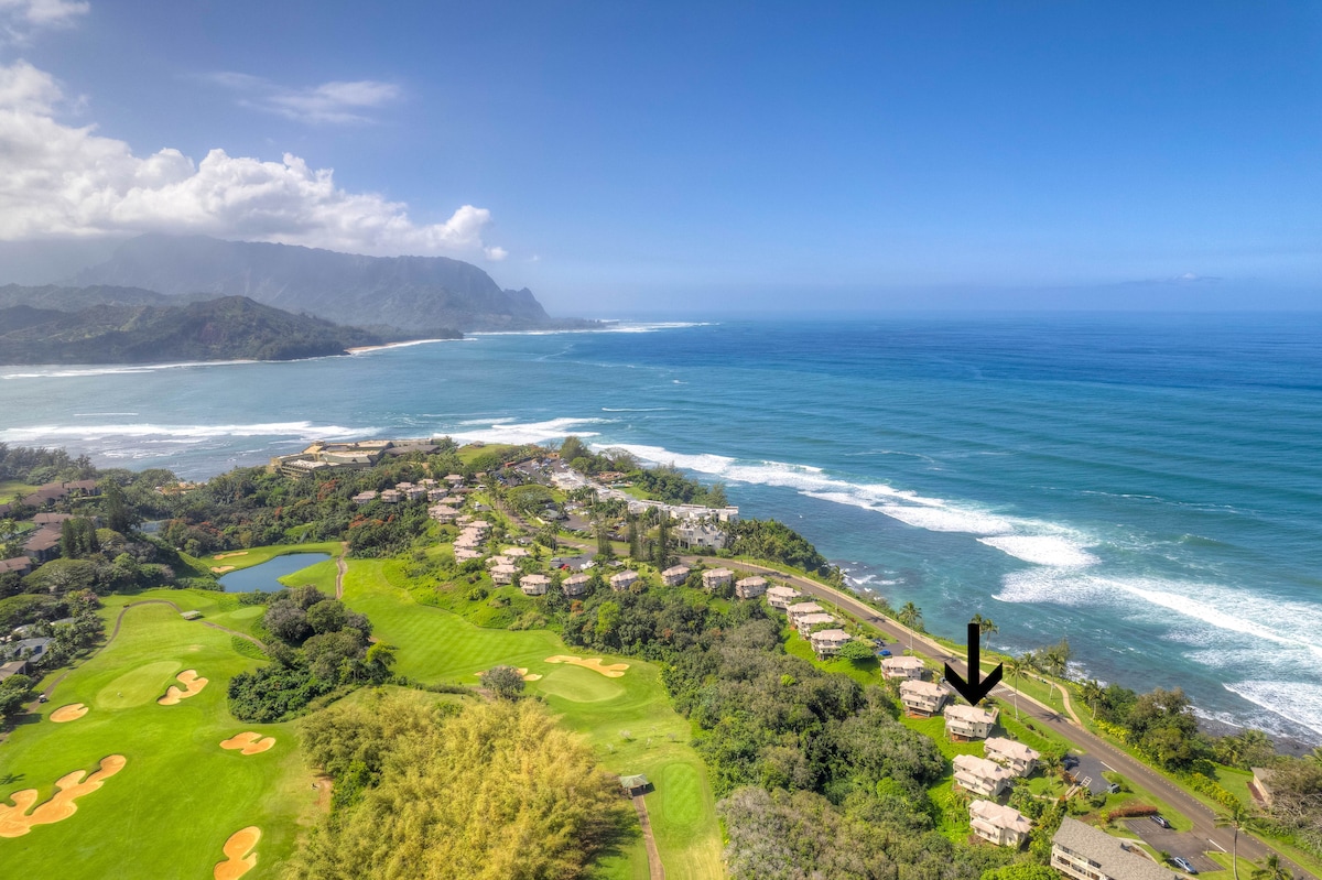 Aerial view of the coastal landscape showcases stunning ocean views and lush greenery. The villa is positioned on a bluff, surrounded by gentle hills and vibrant golf courses, with the beach visible along the shoreline. Waves can be seen breaking along the coast under a clear sky.