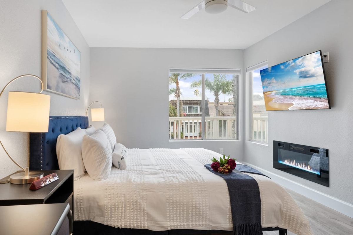 A bright and airy bedroom features a spacious bed with light bedding, complemented by a decorative throw. A wall-mounted smart TV hangs opposite the bed, while an electric fireplace below adds warmth. Large windows offer fresh air and views of palm trees outside.