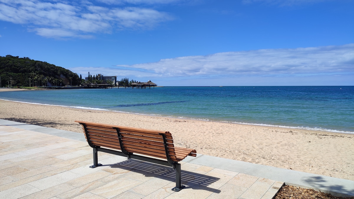 A wooden bench is positioned along a sandy beach, facing the tranquil blue waters. The shoreline is framed by a gentle curve of land, with foliage visible in the background. A clear sky with scattered clouds creates a serene coastal atmosphere.