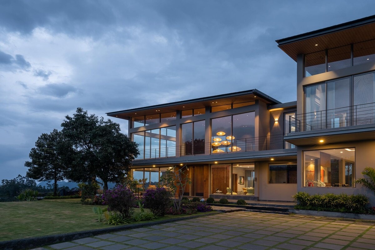 The modern villa is framed by lush greenery, with extensive glass panels reflecting the evening sky. A manicured lawn extends in front, complemented by colorful flower beds, while the exterior features clean lines and warm lighting that creates an inviting atmosphere.