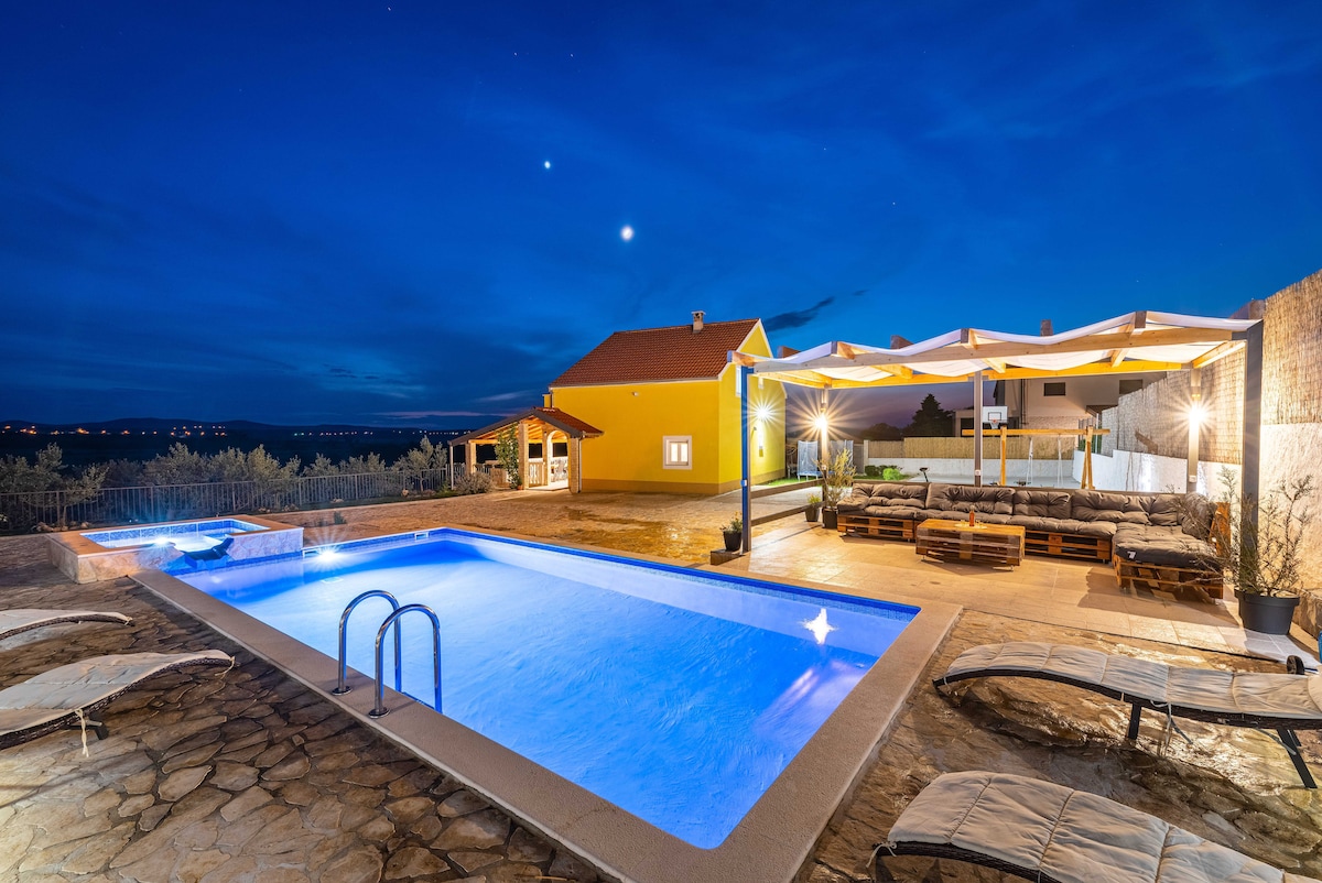 An inviting outdoor area showcases a spacious private swimming pool highlighted by soft blue lighting at night. Comfortable lounge chairs are positioned around the pool, while a shaded seating area features large cushions and a dining table. The surrounding landscape contributes to a relaxing ambiance.