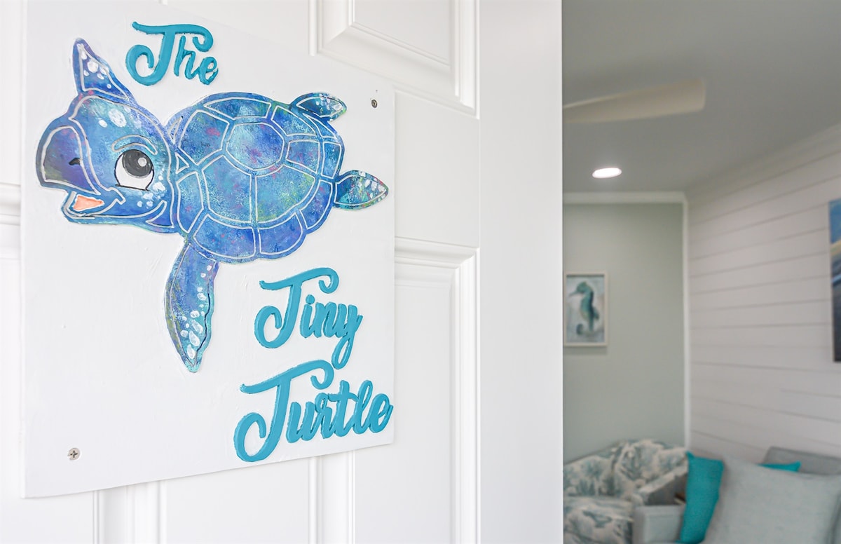 A colorful wall sign featuring a hand-painted turtle design identifies the entrance to the space. The name 'The Tiny Turtle' is displayed in vibrant turquoise, adding a friendly touch to the white door background.