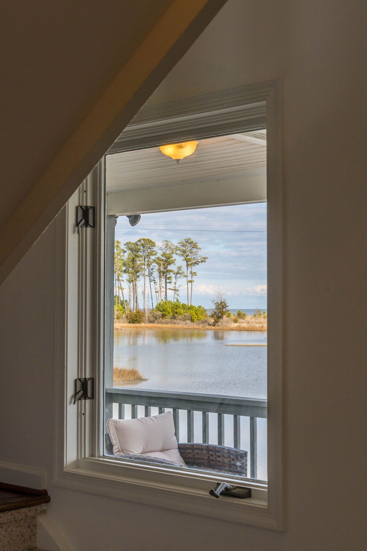 A window frames a serene view of the cove, with calm waters reflecting the sky. Tall trees line the shore, and a cozy seating area with cushions is positioned on the porch, inviting relaxation and scenic enjoyment.