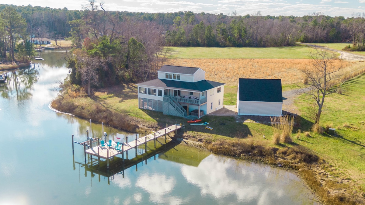 Aerial view of a two-story home situated by a tranquil cove, surrounded by grassy fields and trees. A private dock extends into the water, featuring outdoor seating and kayaks. The serene landscape reflects in the calm water, enhancing the peaceful atmosphere.