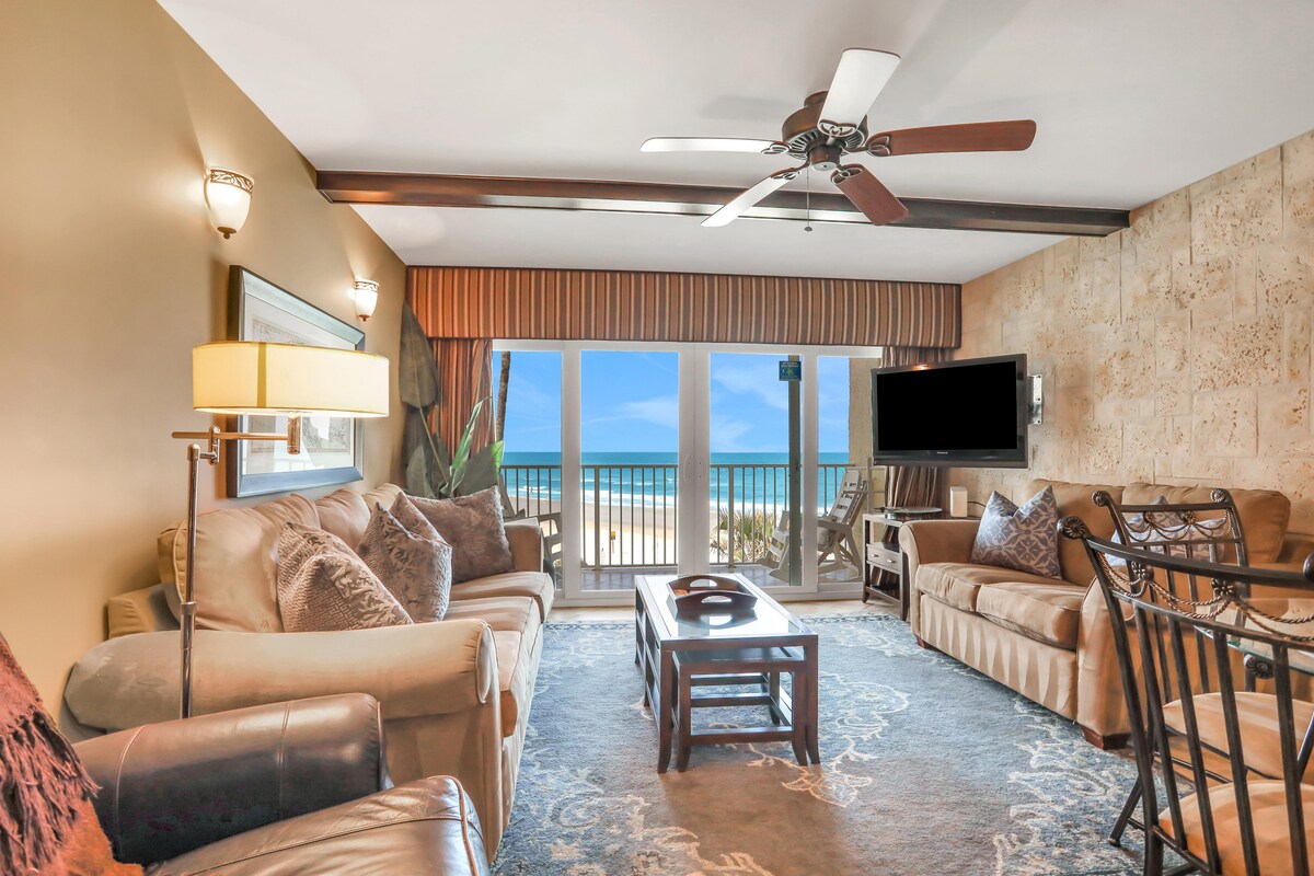 The living area features comfortable seating with a mix of sofas and chairs, a ceiling fan, and a flat-screen television. Large glass doors provide beach views, while the room is adorned with neutral tones and a textured accent wall, creating a relaxed atmosphere.