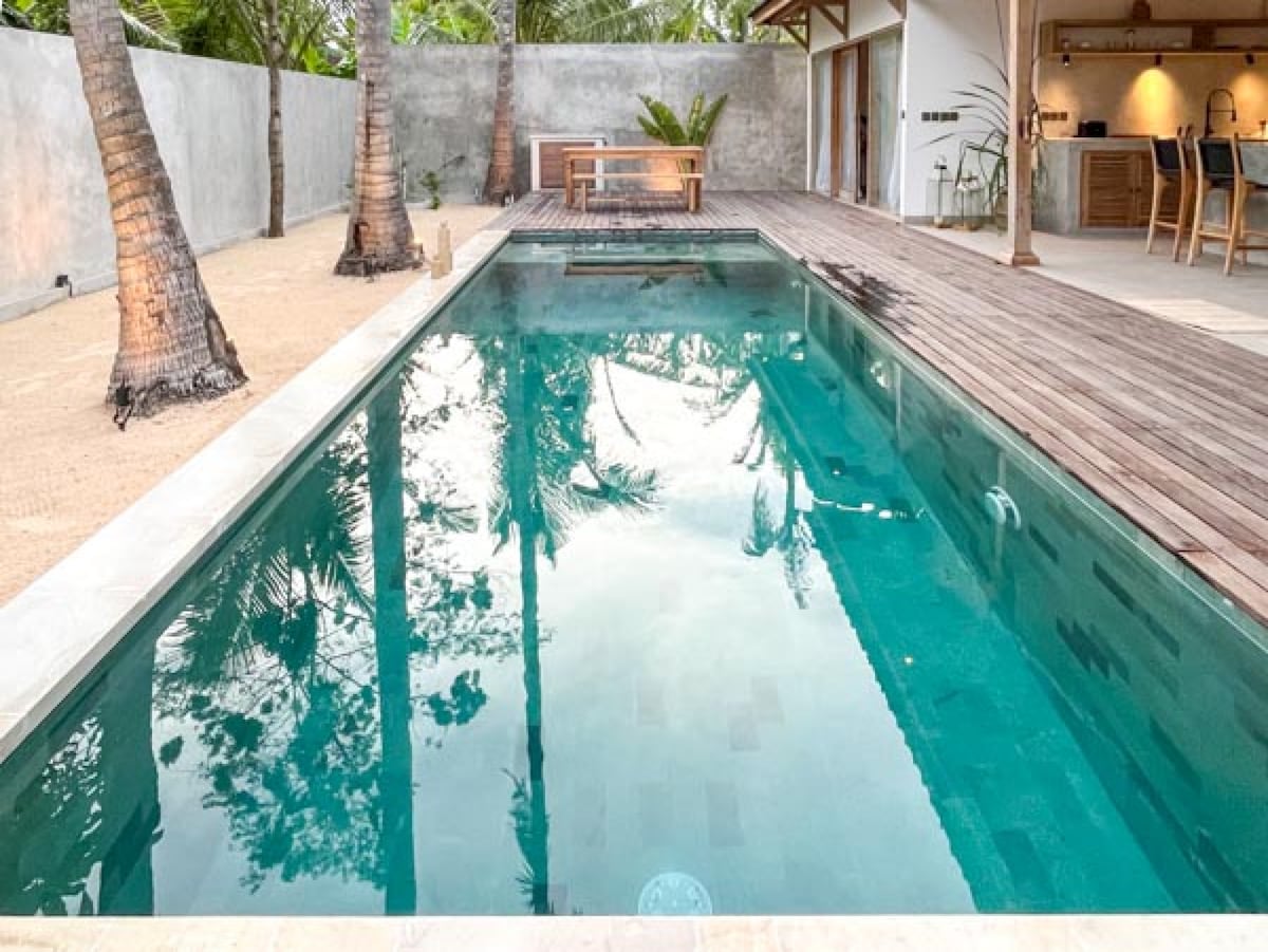 A private swimming pool reflects the surrounding palm trees and sky, surrounded by a wooden deck. A sandy garden area enhances the tropical setting, while a dining table is positioned nearby, inviting relaxation and outdoor meals.