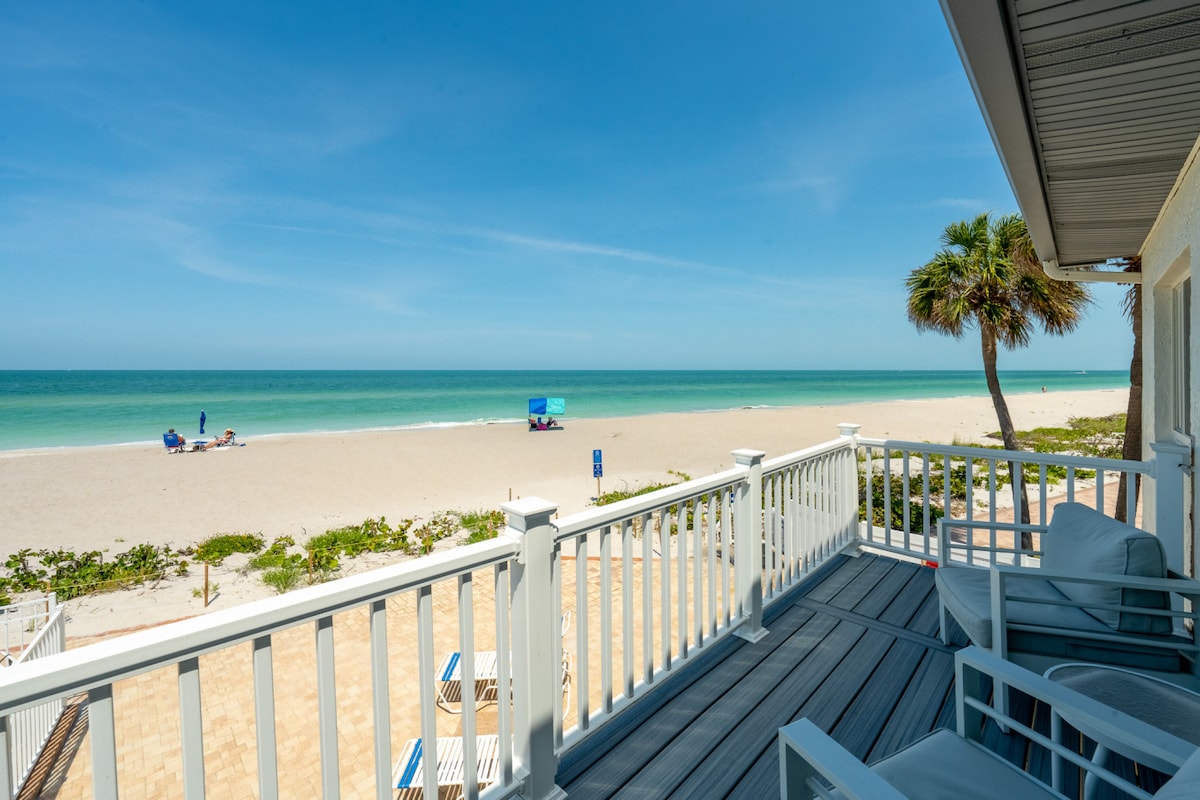 A private balcony offers an unobstructed view of the sandy beach and calm ocean waters. The scene includes lounge chairs positioned for relaxation, with palm trees swaying gently in the breeze. Clear skies and gentle waves enhance the serene ambiance.