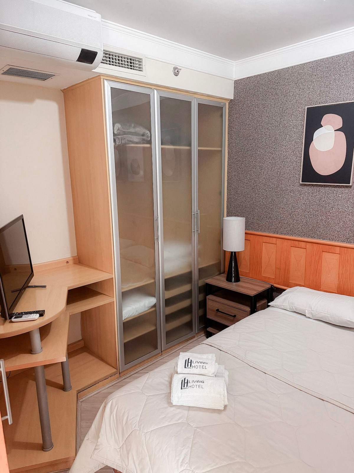 A functional studio space is depicted, featuring a spacious bed with neutral bedding. A compact workstation and a flat-screen television are visible. An organized wardrobe with glass doors stands against the wall, while a nightstand with a lamp completes the room's setup.