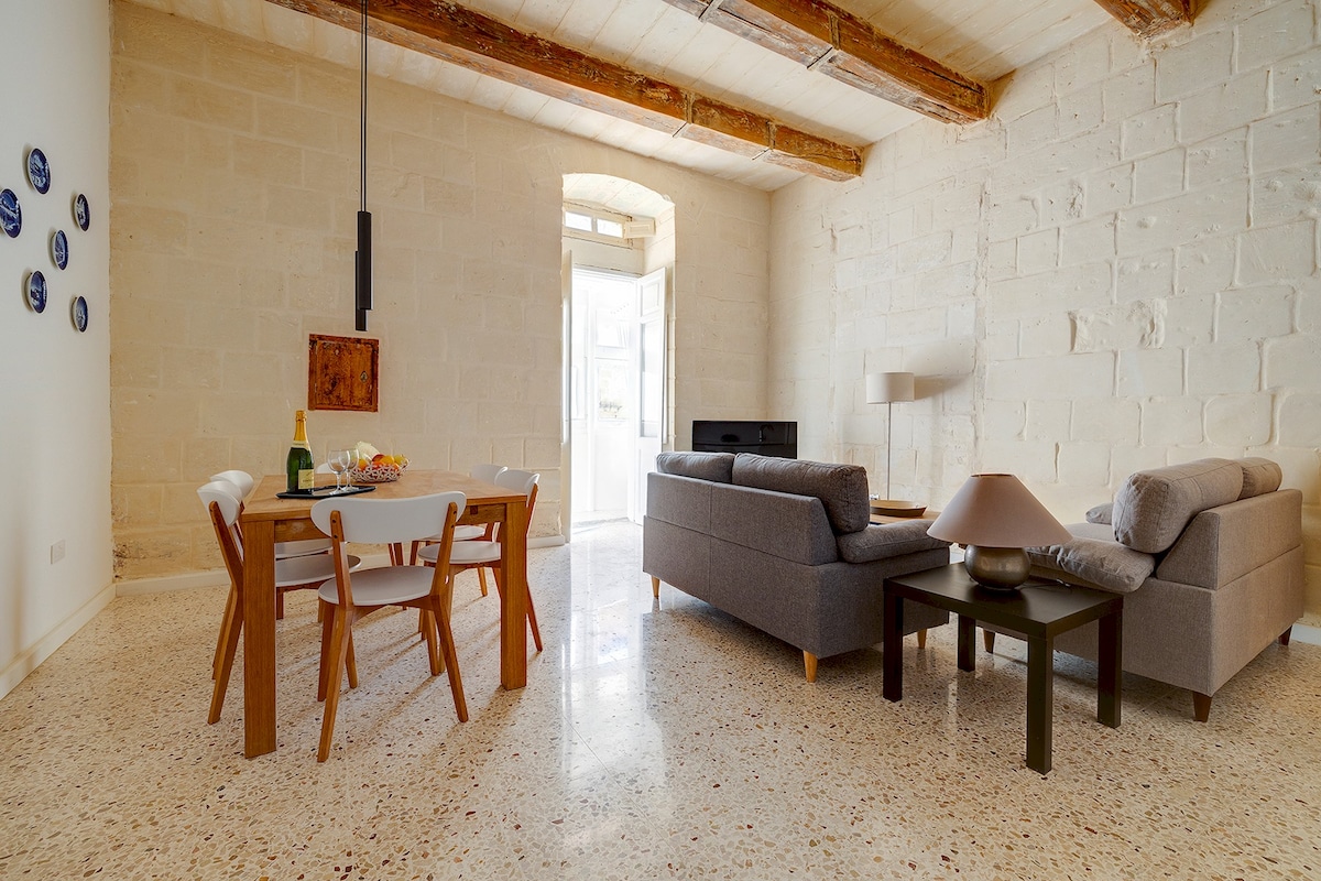 An open-plan living and dining area features a light wood dining table with white chairs and a cozy seating arrangement with grey sofas. Natural light streams in through large doors, illuminating the polished stone floor and highlighting the textured stone walls and wooden beams above.
