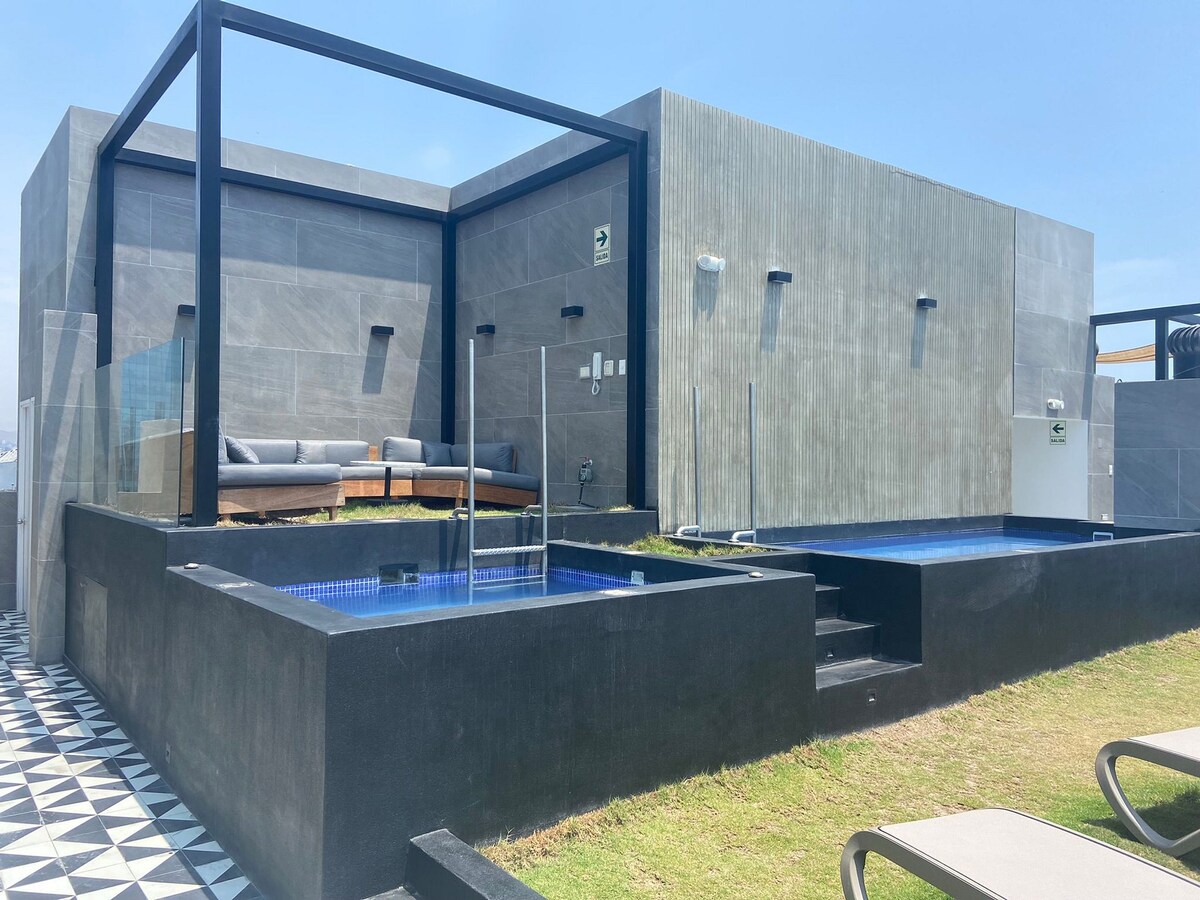 A sunlit rooftop area features two plunge pools surrounded by a sleek black and white tiled deck. Comfortable seating is positioned nearby, creating a relaxing outdoor space for guests. The minimalist design highlights the modern architecture against the clear blue sky.