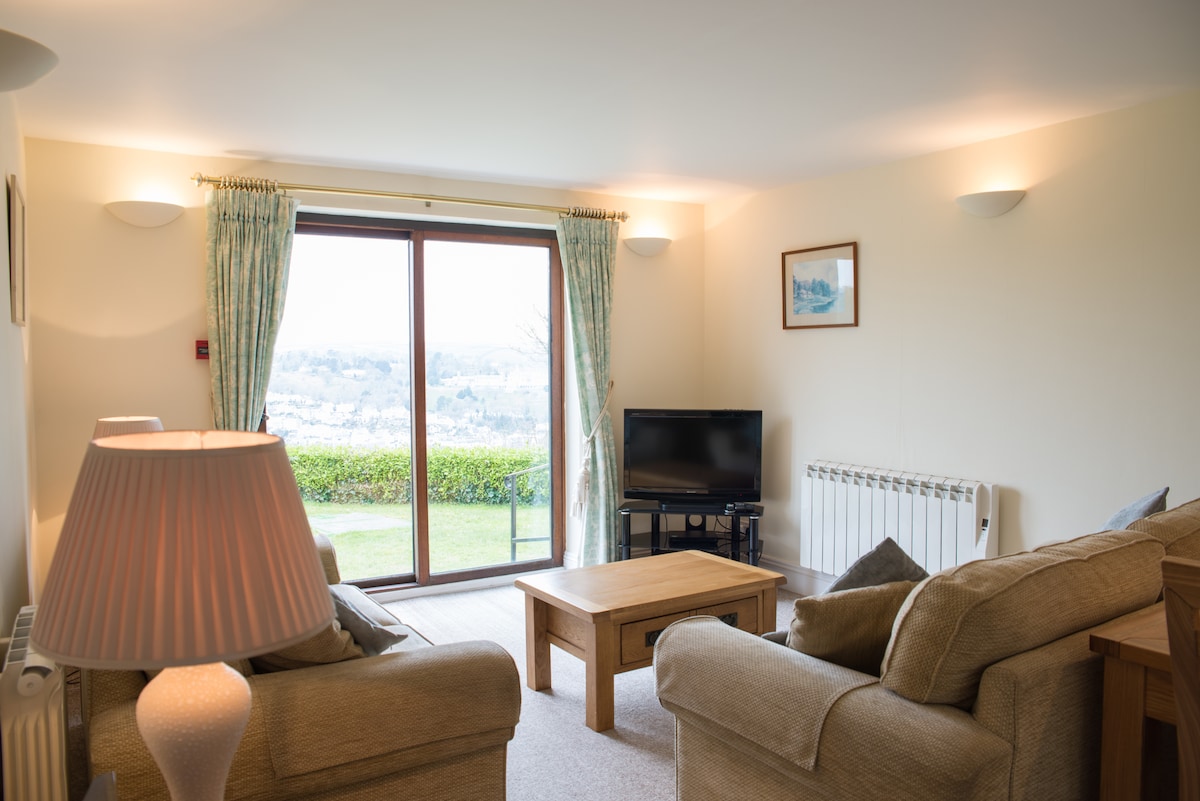 The lounge area features a comfortable seating arrangement, including a spacious sofa and an armchair, positioned around a wooden coffee table. A flat-screen TV is visible, and soft lighting creates a relaxed ambiance. Large patio doors offer views of the lush green landscape outside.