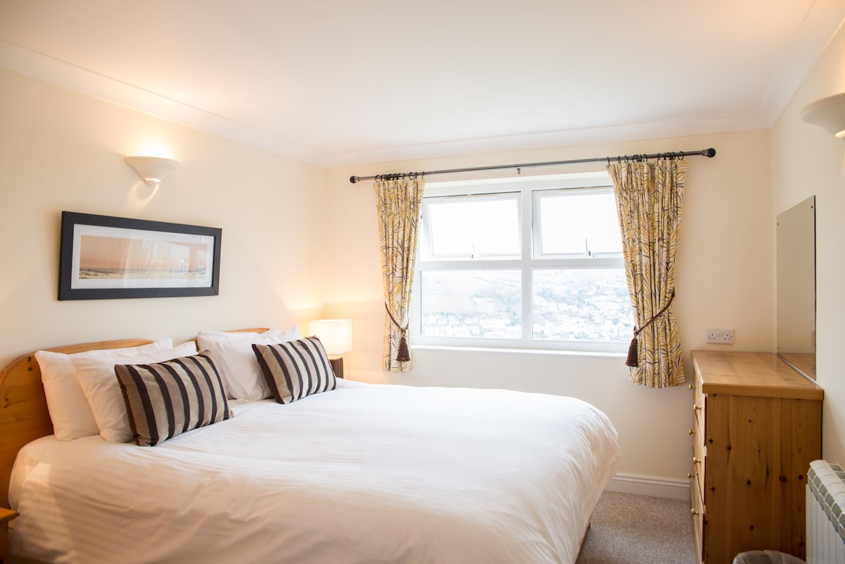 A bright bedroom features a spacious bed with plush pillows and decorative cushions. Natural light fills the room through two large windows with patterned curtains. A wooden dresser is positioned against the wall, accentuating a serene atmosphere.