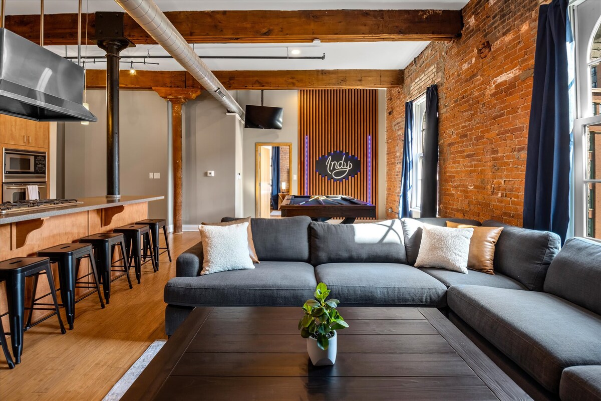 A spacious living area features exposed brick walls and wooden beams, complemented by large windows with dark curtains. A comfortable sectional sofa surrounds a coffee table, while a kitchen island with barstools adds a modern touch, and artwork above the bed enhances the room's character.