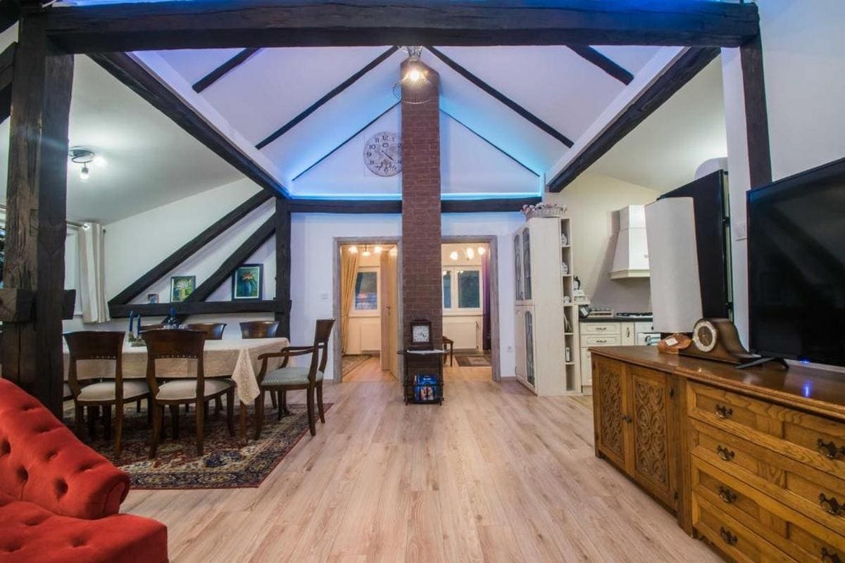 An open-plan living area features exposed wooden beams and a cozy dining space with a wooden table surrounded by chairs. A large television sits opposite a rustic wooden cabinet, while a warm glow illuminates the room from overhead and a central fireplace.