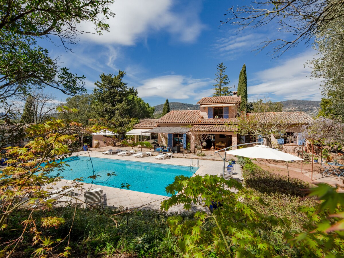 The property showcases a well-maintained swimming pool surrounded by sun loungers and umbrellas. A two-story house with a tiled roof and wooden accents is visible in the background, framed by trees and greenery under a partly cloudy sky.
