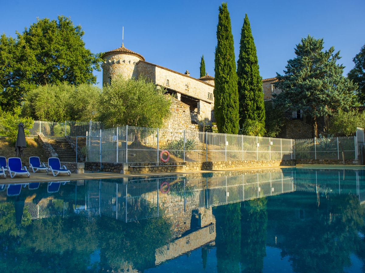 A stone residence is reflected in the calm water of a large swimming pool. Tall trees frame the property, and a sun terrace with blue loungers is visible. The scene is set under a clear blue sky, capturing the peaceful ambiance of the countryside.