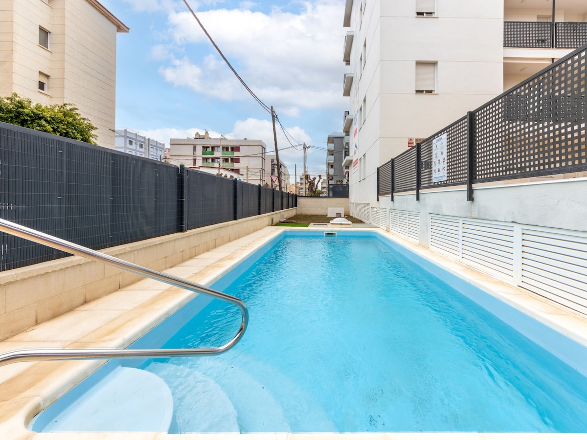 A rectangular swimming pool is featured within a fenced area, with clear blue water inviting relaxation. Light-colored pool tiling contrasts with the surrounding concrete. The setting is enhanced by nearby buildings and a cloudy sky.