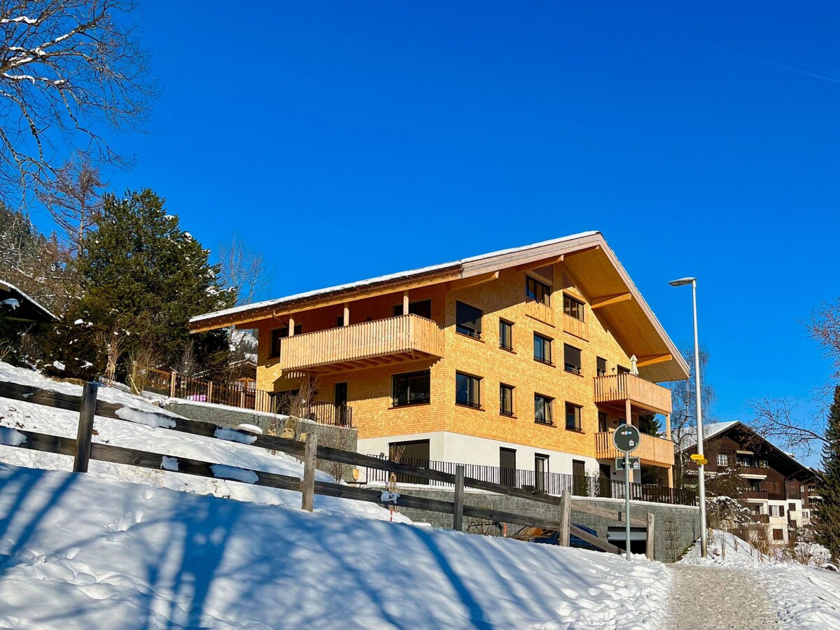 A modern building constructed with light wood is surrounded by snow-covered grounds. The structure features multiple floors, large windows, and balconies. Clear blue skies provide a contrasting backdrop, enhancing the inviting appearance of this elevated holiday flat.