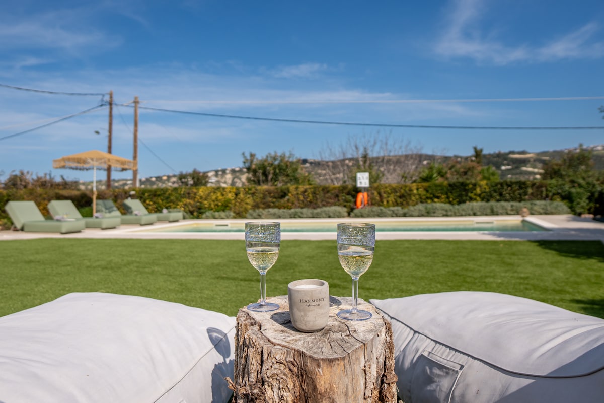 Two glasses of sparkling beverage are placed on a rustic wooden stump between two comfortable lounge chairs. A well-maintained green lawn leads to a private swimming pool, which reflects the clear blue sky and surrounding landscape.