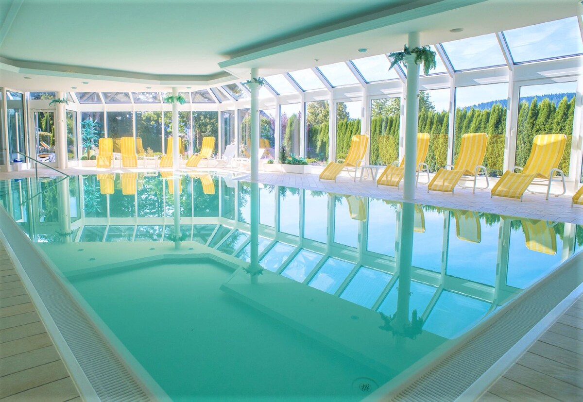 An indoor pool area is surrounded by large glass windows, allowing natural light to flood the space. Yellow lounge chairs are positioned around the pool, reflecting off the clear water. Lush greenery can be seen outside, adding a serene backdrop.