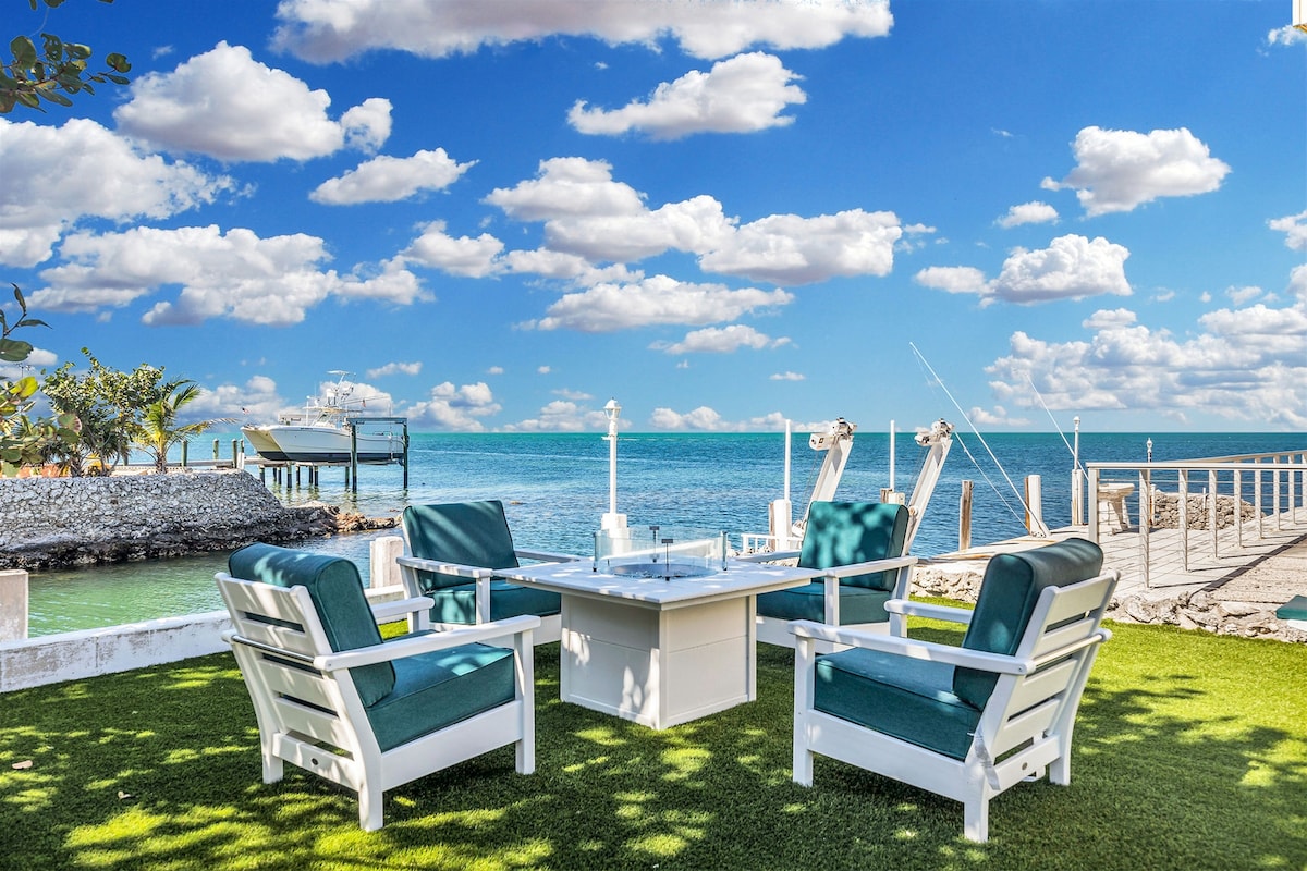 Outdoor seating area is featured along the waterfront, consisting of four white lounge chairs surrounding a fire pit. The vibrant green grass enhances the setting, while the blue sky is dotted with fluffy clouds above the calm turquoise waters.